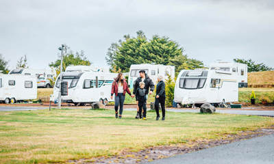 A family of two parents and two children wearing coats and hats walking on a green field in front of touring vans and motorhomes 