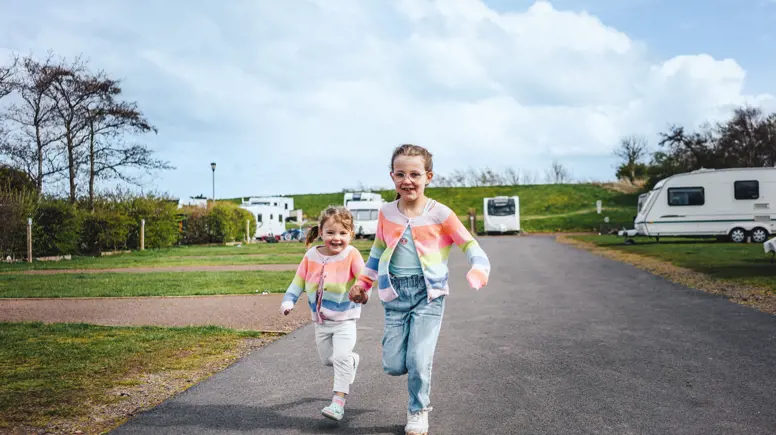 Two little girls in matching colourful cardigans holding hands and running next to touring caravans on pitches 