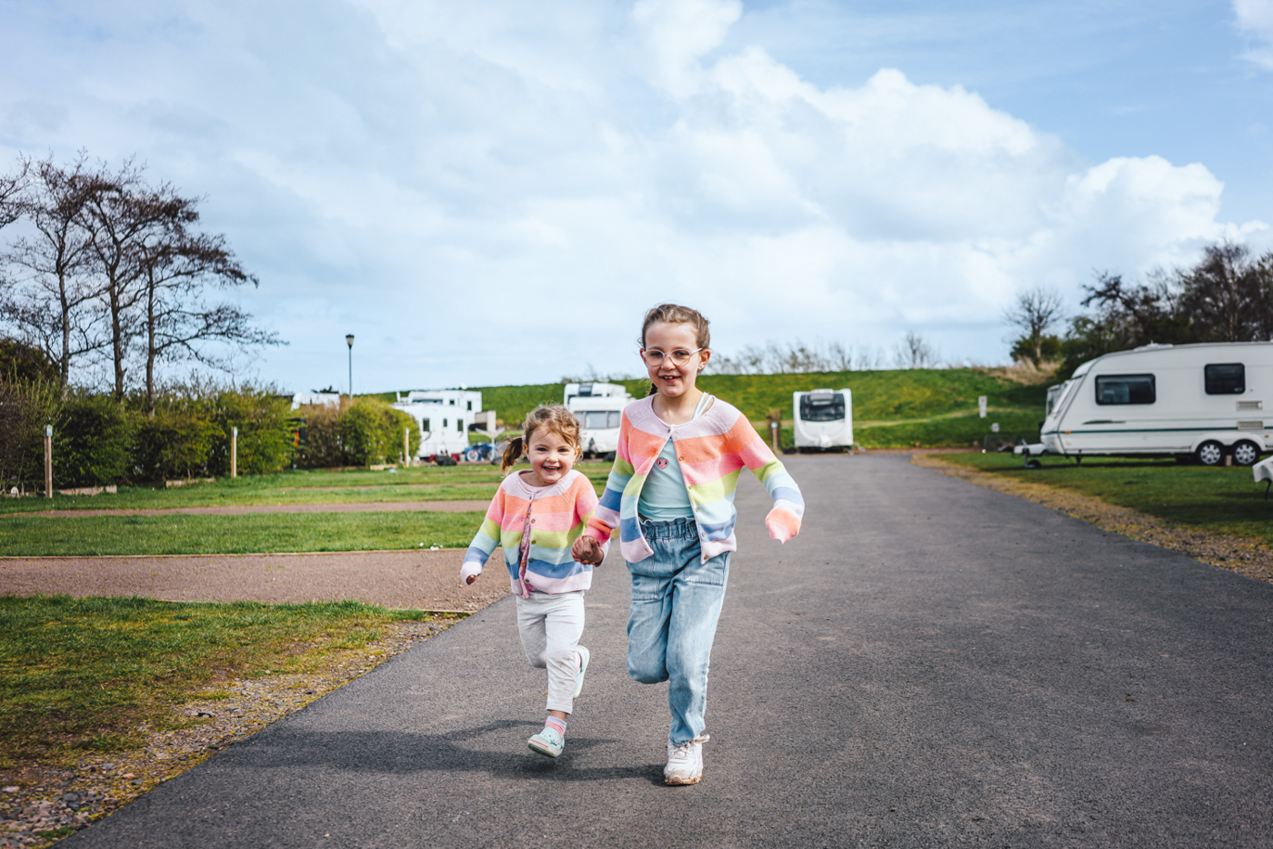 Two little girls in matching colourful cardigans holding hands and running next to touring caravans on pitches 
