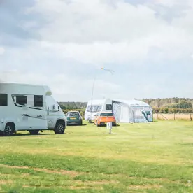 Two touring vans and cars parked on touring pitches at a UK holiday park with green grass and a blue sky above 